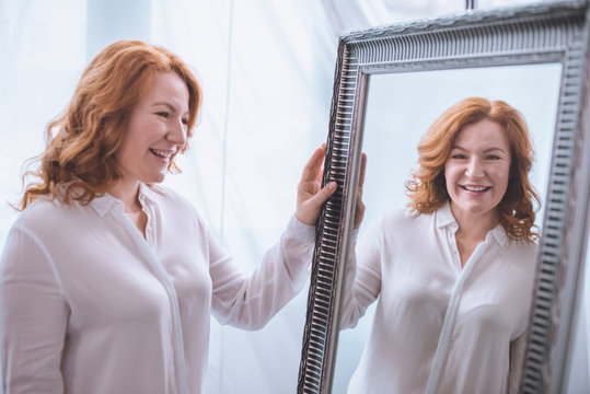 beautiful smiling mature woman standing near mirror and looking at reflection