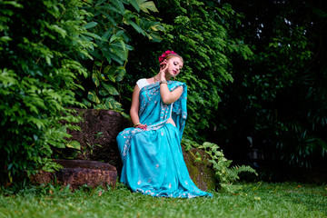 Obraz na płótnie Canvas Beautiful young caucasian woman in traditional indian clothing sari with bridal makeup and jewelry and henna tattoo on hands sitting in green garden.