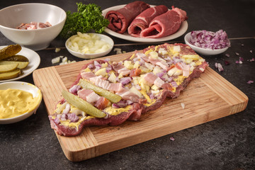 preparing beef roulades on a wooden cutting board, fresh raw meat with mustard, onions, pickled cucumber and bacon, dark kitchen countertop