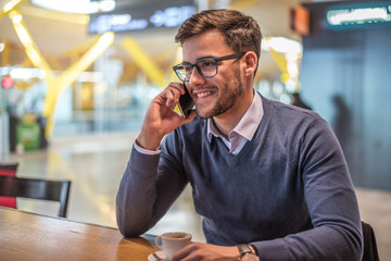 young man at the airport using his mobile phone and drinking coffee waiting for his flight close-up portrait