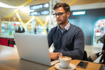 young man at the airport working with laptop using his mobile phone and drinking coffee waiting for...
