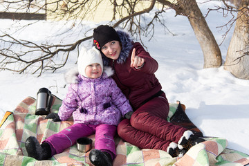 Fototapeta na wymiar Mom and daughter are sitting on a plaid in the snow in the winter. A young mother shows her little daughter the sky.