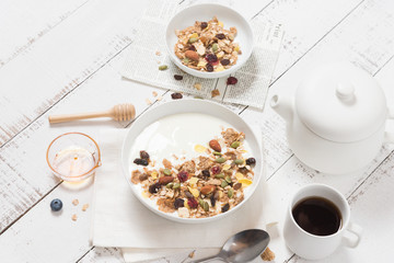 Cereal breakfast with dried almond, cranberry, grapes, Pistachios nuts and cornflakes over plain yoghurt in white bowl with honey, black coffee cup and spoon on white wooden background, healthy meal