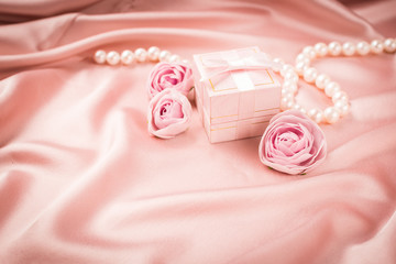 A gift in a pink box is tied with a satin ribbon necklace of pearls and flowers on a background of a delicate pink satin. Copy space.
