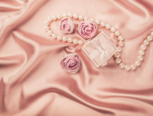 A gift in a pink box is tied with a satin ribbon necklace of pearls and flowers on a background of a delicate pink satin. Holiday concept. Flat layout.