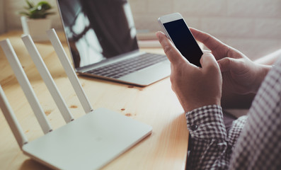 Mockup image of hand holding white mobile phone with blank black screen and wifi router on vintage wood table in home