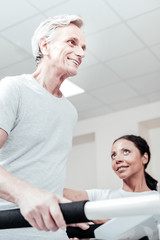 Obraz premium Recovery. Exuberant energetic old grey-haired man smiling and exercising on a training device and a smiling dark-haired dark-eyed woman sitting near him and looking at him