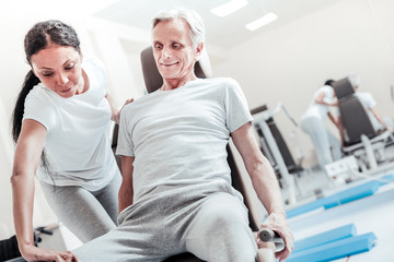 Obraz premium Therapist. Gleeful determined old grey-haired man smiling and exercising on a training device while a dark-haired dark-eyed afro-american woman helping him