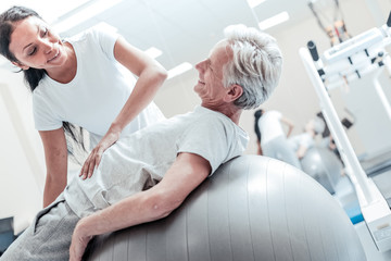 My favourite trainer. Exuberant old wrinkled grey-haired man lying on a ball for exercises and a joyful young smiling dark-haired afro-american woman helping him and touching his stomach