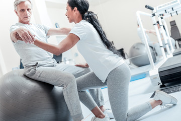 Fototapeta na wymiar Exercises. Concentrated old grey-haired man sitting on a ball for exercises and a young smiling dark-haired afro-american woman helping him to train his arms and looking at him