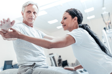 Fototapeta na wymiar Do like this. Concentrated old grey-haired man sitting on a ball for exercises and a beautiful young dark-haired afro-american woman helping him to train his arms and touching his arm and leg