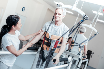Learning to walk. Vigorous old grey-haired man smiling and exercising on a training device and learning to walk and a young dark-haired female trainer standing near him and touching his hand