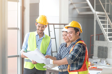 Three Asian male and female architect engineers with helmeted smiling and talking together while reading plan in paper standing at construction site background.