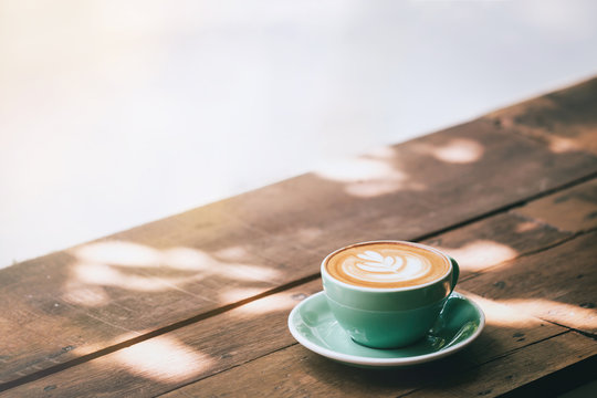 Fototapeta Hot cappuccino coffee in a green cup with latte art on the wooden table in a coffee shop and glass background. Vintage toned.
