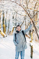 Young man in gray jacket and blue jeans with a black backpack walking in the winter park