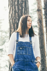 Beautiful young woman in jeans overalls standing in woodland