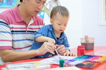Fototapeta premium Cute little Asian 18 months / 1 year old toddler baby boy child painting with brush and watercolors, kid painting with father at home ,Creative play for toddlers concept