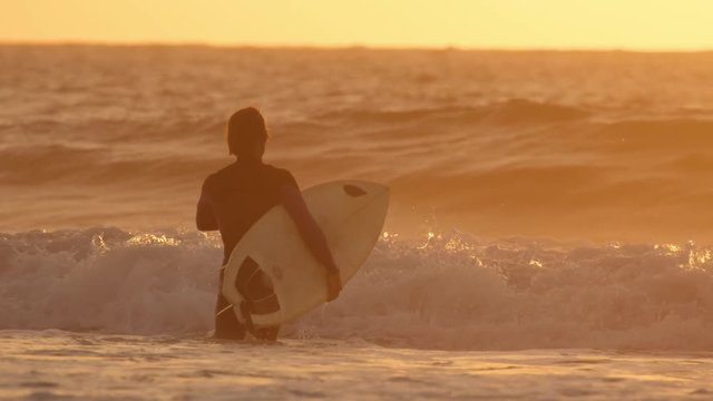 SLOW MOTION: Unrecognizable surfer in black wetsuit going surfing big crashing waves at epic sunset. Easygoing surfer dude walking in deep ocean water to go surf glassy ocean waves before nightfall.