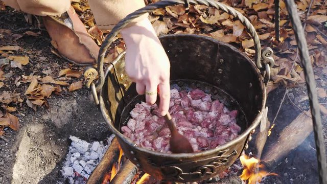 Close-up of a woman's hand prevents meat in a cauldron on an open fire in forest