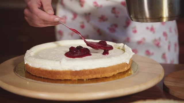 Housewife is covering layer of cake with butter cream by berries jam. She is stirring jam in a cup and taking by spoon, close-up