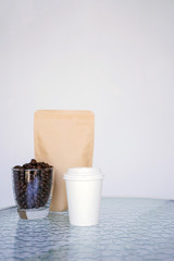 A white paper glass (one-time) for coffee and coffee beans in transparent glass with paper craft container is on the table. Background is blurred, white and blue. Place for the title on the glass.
