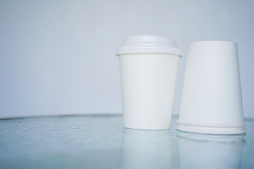 Two white paper glass (one-time) for coffee is on the table. Background is blurred, white and blue. Place for the title on the glass. Side view with copy space