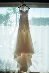 white bride dress hang againt big door or window on white curtain with sunlight