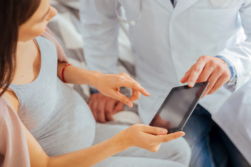 Innovative medicine. Top view on an expectant mother and pediatrician sitting next to each other and talking while looking at a screen of a tablet computer.