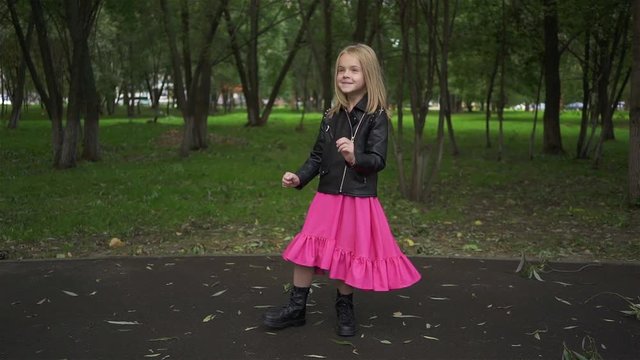 Cute little blonde girl wearing a leather jacket and a pink dress is dancing while standing in a park on a summer day. Handheld slow motion medium shot