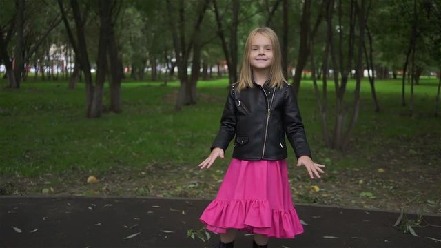 Cute little blonde girl wearing a leather jacket and a pink dress listening to the music standing in a park on a summer day. Zoom out real time medium shot