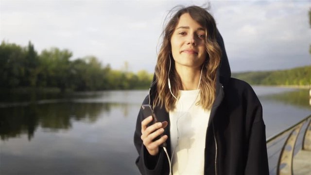 Positive young woman wearing a black hoodie listening to the music and dancing on a river bank. Left to right pan slow motion establishing shot