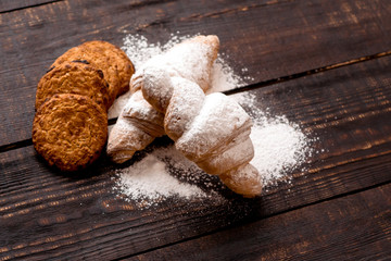 croissants sprinkled with powdered sugar and oatmeal cookies on a dark wooden table