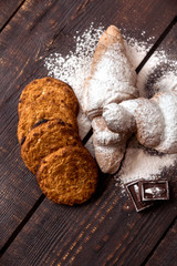 croissants sprinkled with powdered sugar and oatmeal cookies on a dark wooden table