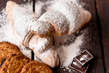 croissants sprinkled with powdered sugar and oatmeal cookies on a dark wooden table
