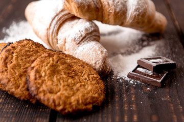 croissants sprinkled with powdered sugar and oatmeal cookies on a dark wooden table