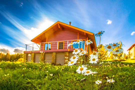Cottage In The Background Of A Summer Day. Wooden Cottage. Flowers On The Background Of The House. Private House With Balcony.