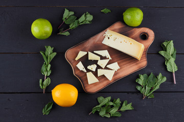 Hard yellow cheese, lemon, lime and fresh mint on a wooden black background, slices of cheese on a black background, top view, mint leaves with cheese with holes, rustic style, citrus, healthy food, v