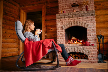 Cozy home. Pretty young woman drinking hot  beverage near the fireplace.