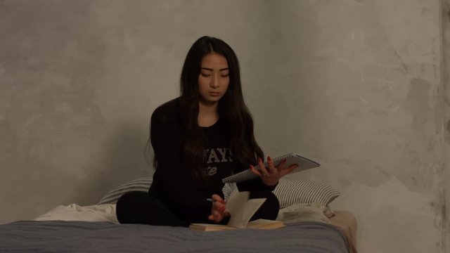 Frustrated charming asian girl throwing textbook away while sitting cross legged on the bed in bedroom and studying hard for college exam. Exhausted female student tired of learning at home.