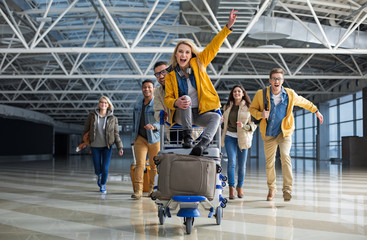 Excited male and female people running to departure gate. One of them is wheeling baggage cart with woman sitting on the top