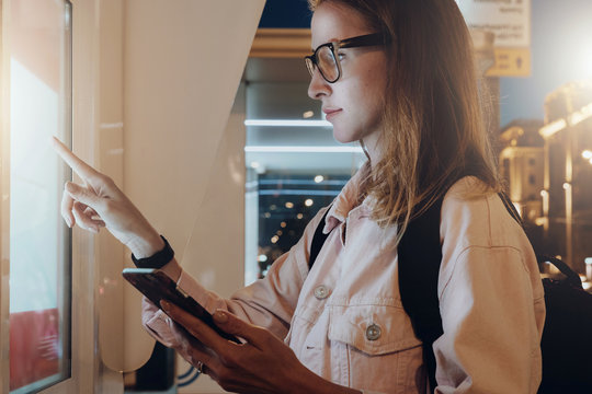 Summer night. Young woman in eyeglasses with backpack stands on city street, touches digital display, holding smartphone in her hand. Innovative technologies, digital display, urban navigation.Mock up