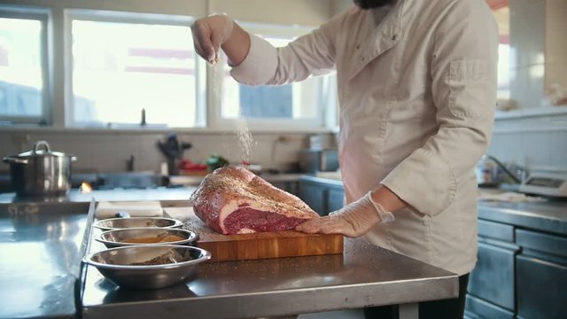 Butcher adding spices into large piece of fresh raw meat lying on a wooden board in a commercial kitchen, slow motion