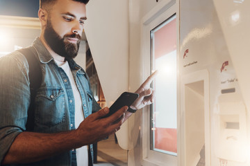 Summer night. Young hipster man with beard stands on city street and touches digital display while...