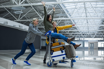Fototapeta na wymiar Delighted women entertaining at the airport. They are carrying luggage trolley with girl sitting on the top of bags