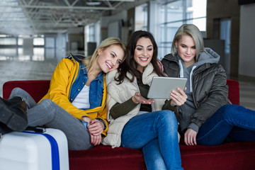 Happy female friends waiting for boarding at the airport lounge. They are sitting on sofa and using tab