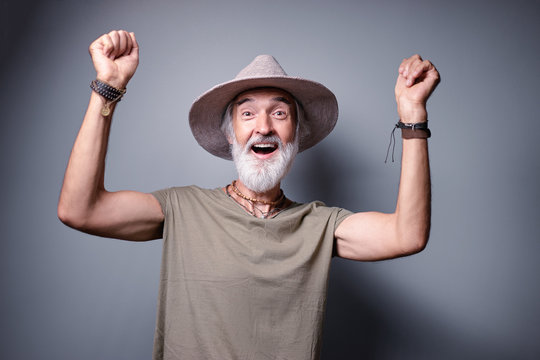 Winner! Studio portrait of excited senior man with gray beard and hat rising hands up.