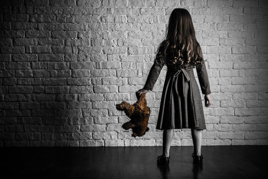 A girl with long hair stands by a white brick wall. In hand soft toy bear.
