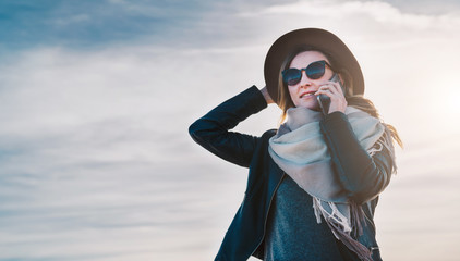 Fototapeta premium Young woman tourist in sunglasses and hat stands outdoor and talks on her cell phone. In background sky, clouds. Girl is calling her friend on phone, using digital gadget. Tourism, trip, vacation.