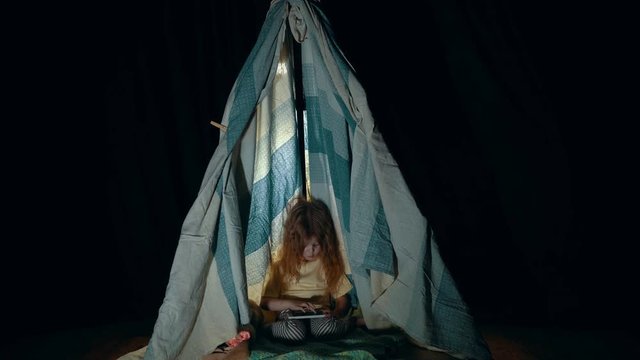A little girl in pajamas uses the phone while sitting in a tepee tent in the playroom in the evening.