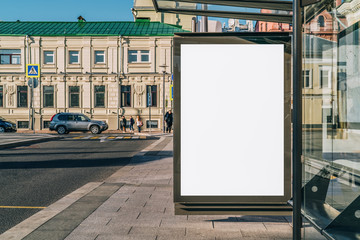 Obraz premium Pionowy pusty biały billboard na przystanku autobusowym na ulicy miasta. W tle budynki i droga. Makieta. Plakat na ulicy obok jezdni. Słoneczny letni dzień.
